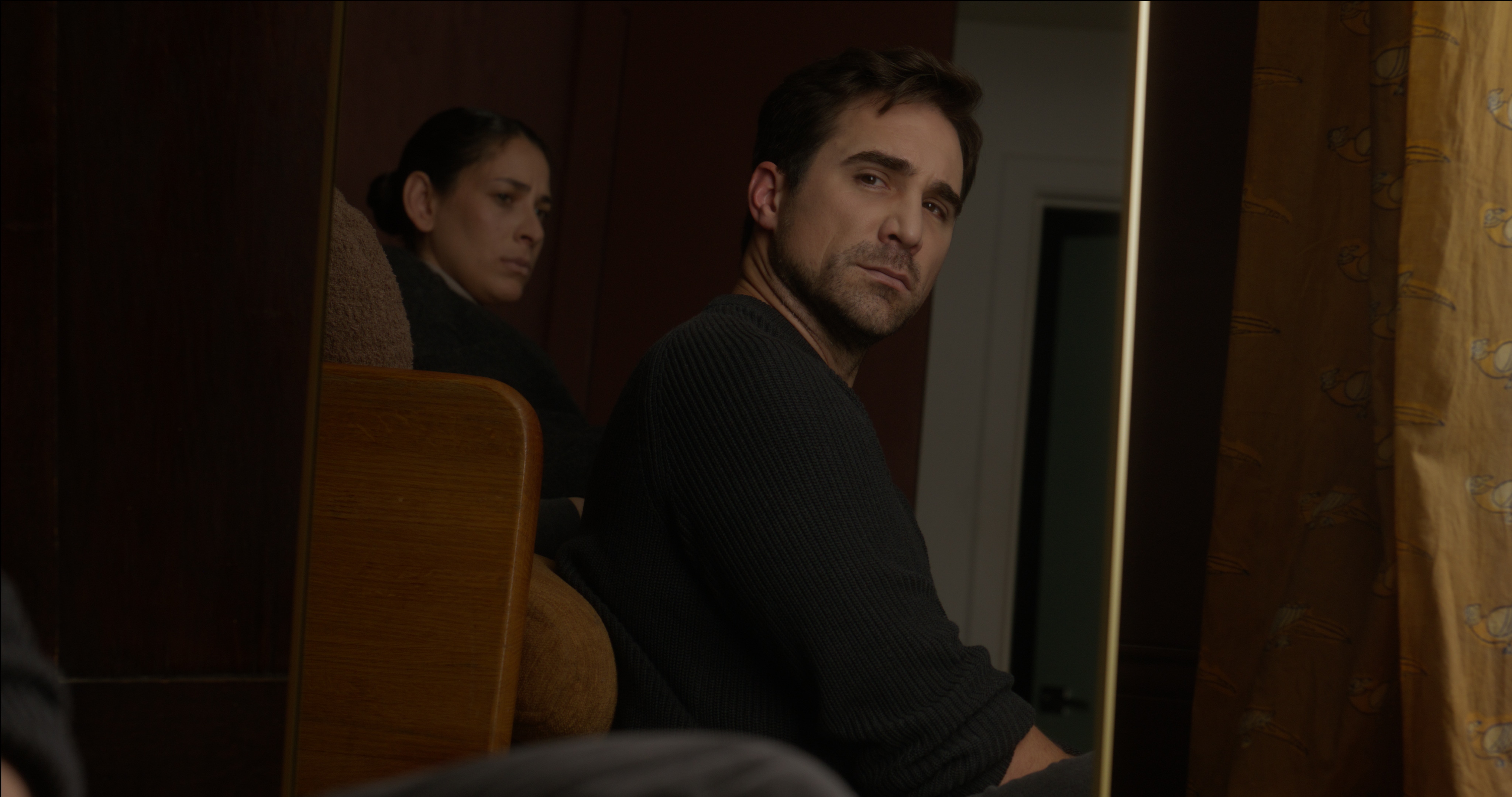 Still from The Conditions of Being Seen. A man glances back over his shoulder while a woman is reflected in a mirror behind him.