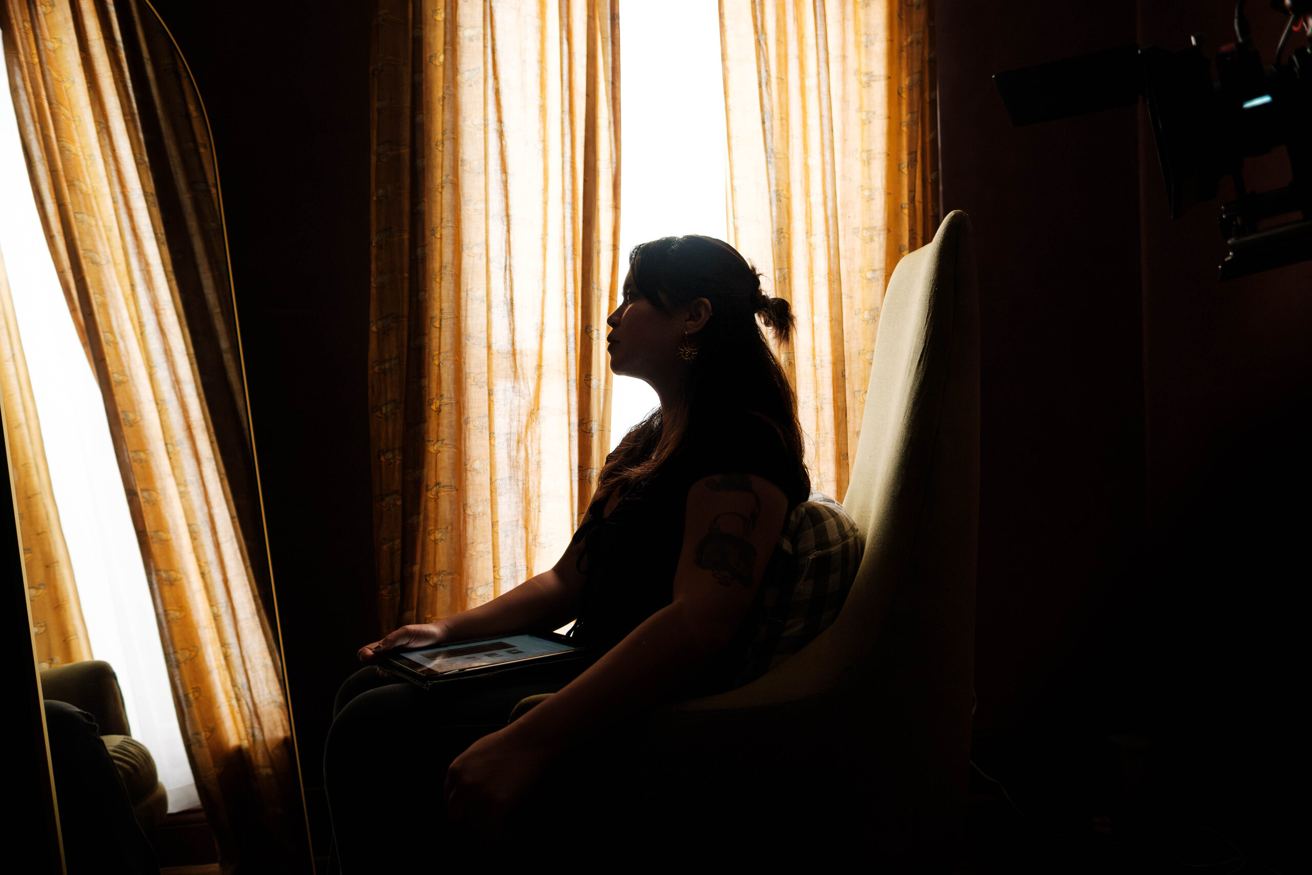 A crew member sits silhouetted against the curtained window beside the camera.