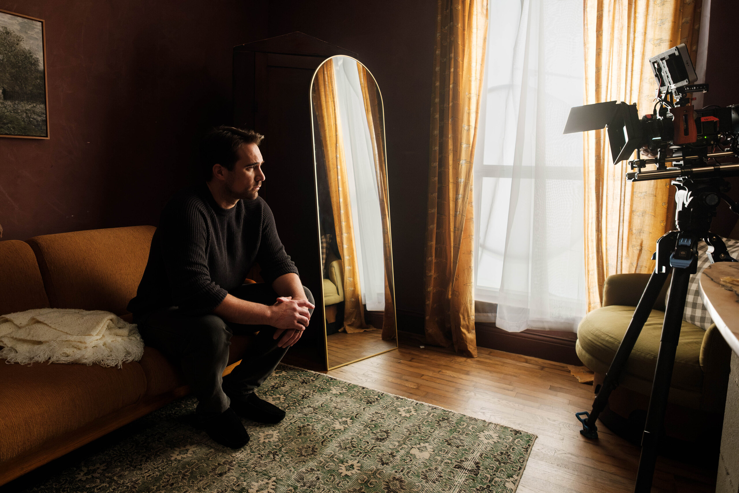 Justin Ray sits on the sofa on set, the camera standing by.
